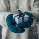 a person wearing a cream winter sweater and blue mittens holding a small gift wrapped in white paper and blue ribbon in outstretched hands. it is snowing