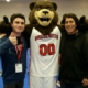a college student gives a thumbs up with right hand while left arm is around his college mascot, a bear in a basketball uniform, his father stands on the other side of the mascot