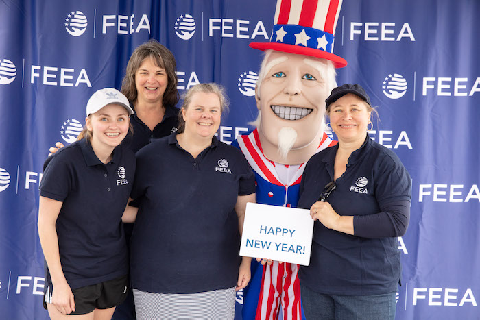 FEEA staff Alyssa, Niki, Robyn, and Joyce wearing dark blue polo shirts with FEEA logos stand with a person in Uncle Sam costume with red, white, and blue tophat in front of a blue step-and-repeat of the FEEA logo holding a Happy New Year sign