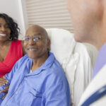 Happy senior African American man patient recovering in hospital bed with male doctor and wife