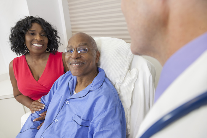 Happy senior African American man patient recovering in hospital bed with male doctor and wife as his patient advocate.