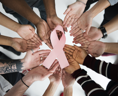 a circle of hands of all races holding a pink breast cancer ribbon in the middle