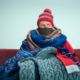 Bearded Man bundled in many scarves and blankets Sitting on Sofa at home wondering how to he can stay warm and be proactive in controlling heating costs
