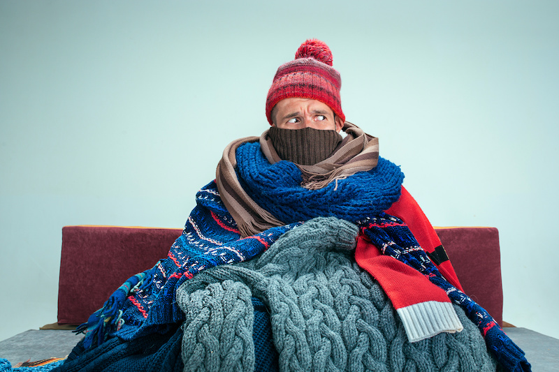 Bearded Man bundled in many scarves and blankets Sitting on Sofa at Home.