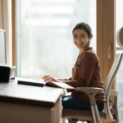 A woman sits at her desk smiling for the camera. Her ergonomic workspace setup includes ergonomic equipment and good posture that plays a role in occupational injuries prevention.