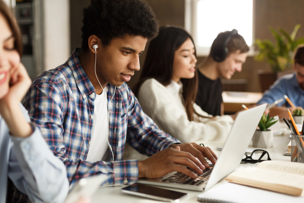 A student participates in FEEA’s Online Tutoring program from his computer with headphones in while sitting around other students working at their computers.