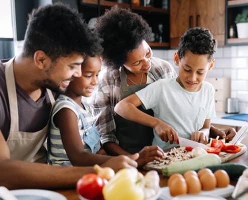 An African American family with a woman and man standing behind a boy and girl smiling as they prepare food in the kitchen.