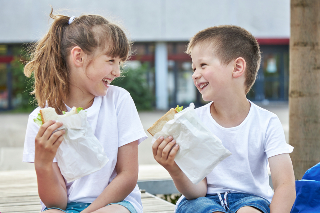 Two kids smile at each other as they hold their sandwiches at lunch. Our expert shares her tips for creating healthy school lunches they'll love