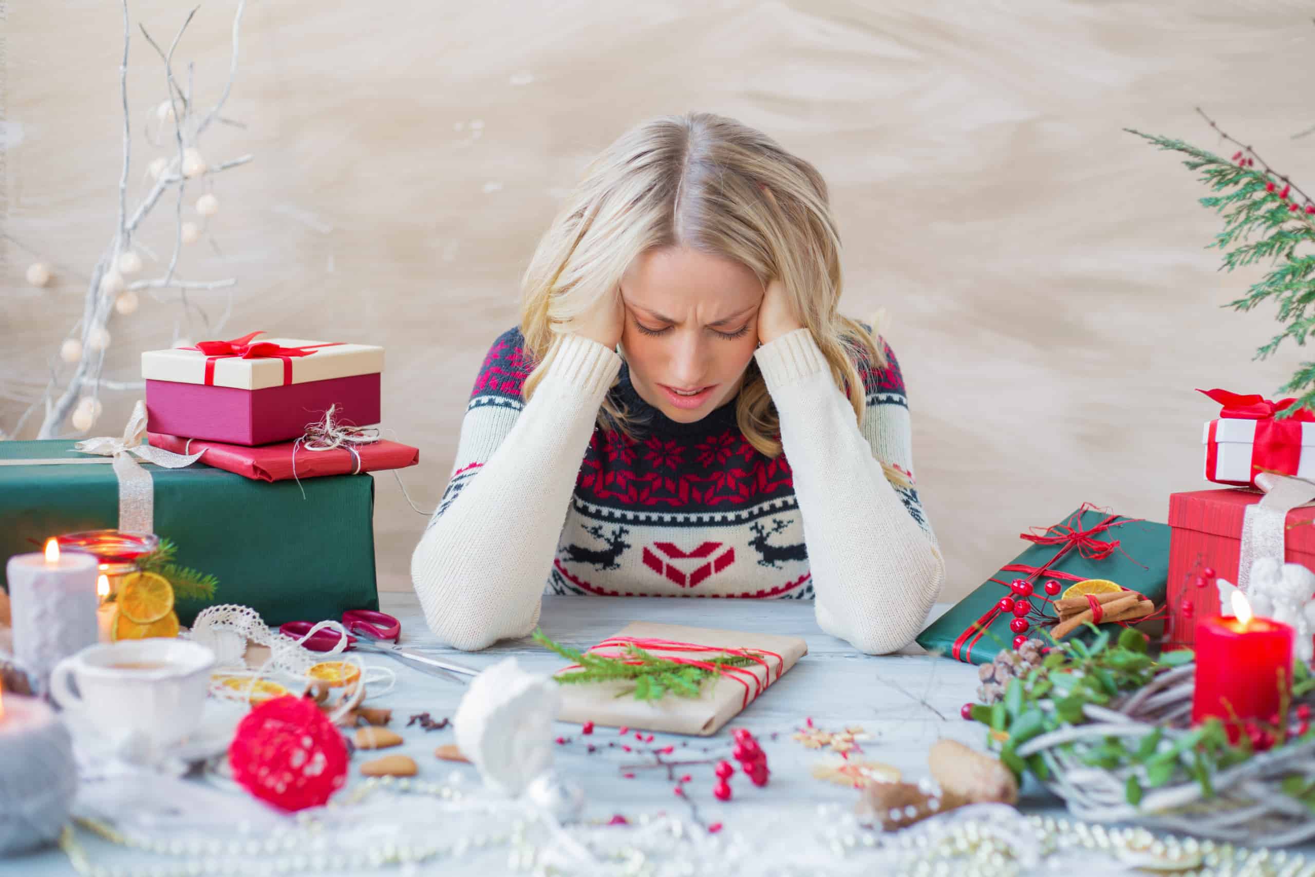 A blonde woman wearing a winter holiday sweater surrounded by presents and other holiday decorations sits at a table with her hands to her head to represent that she is stressed out.