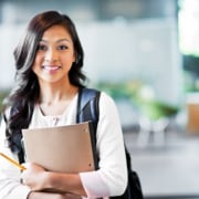 An Asian American woman smiling at the camera, holding a notebook and wearing a backpack. A number of scholarships are available through FEEA, including a first-generation scholarship.
