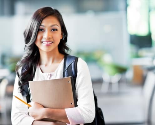 An Asian American woman smiling at the camera, holding a notebook and wearing a backpack. A number of scholarships are available through FEEA, including a first-generation scholarship.