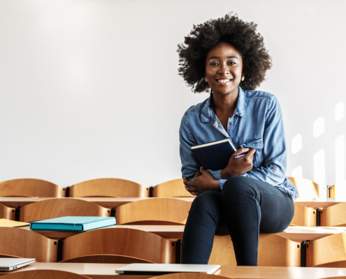 An African American woman smiles for the camera while sitting atop desks in a classroom. FEEA has compiled the best resources to guide students and parents through the college admissions journey.