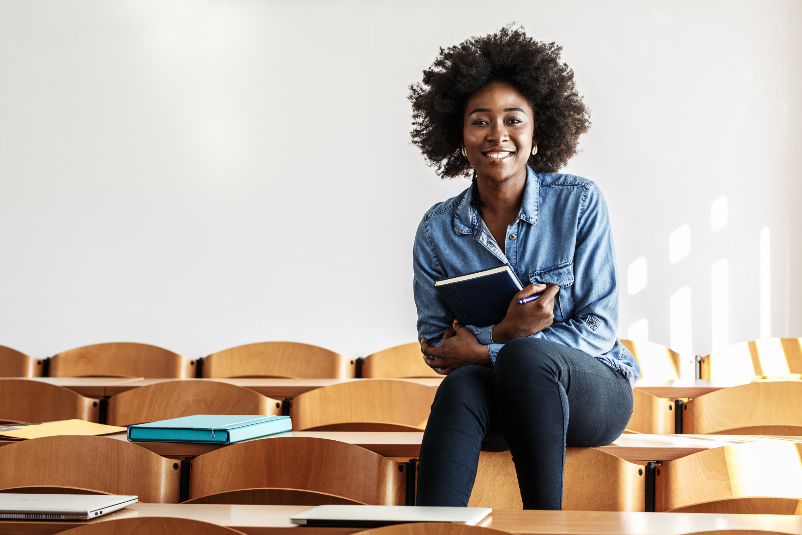 An African American woman smiles for the camera while sitting atop desks in a classroom. FEEA has compiled the best resources to guide students and parents through the college admissions journey.