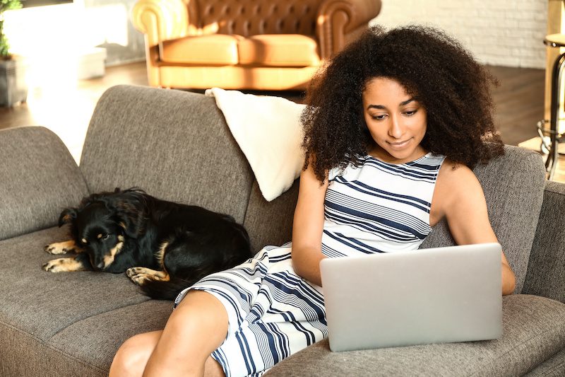 An African American woman sits on the couch next to her dog as she reads through applications as a virtual volunteer of the scholarship selection committee
