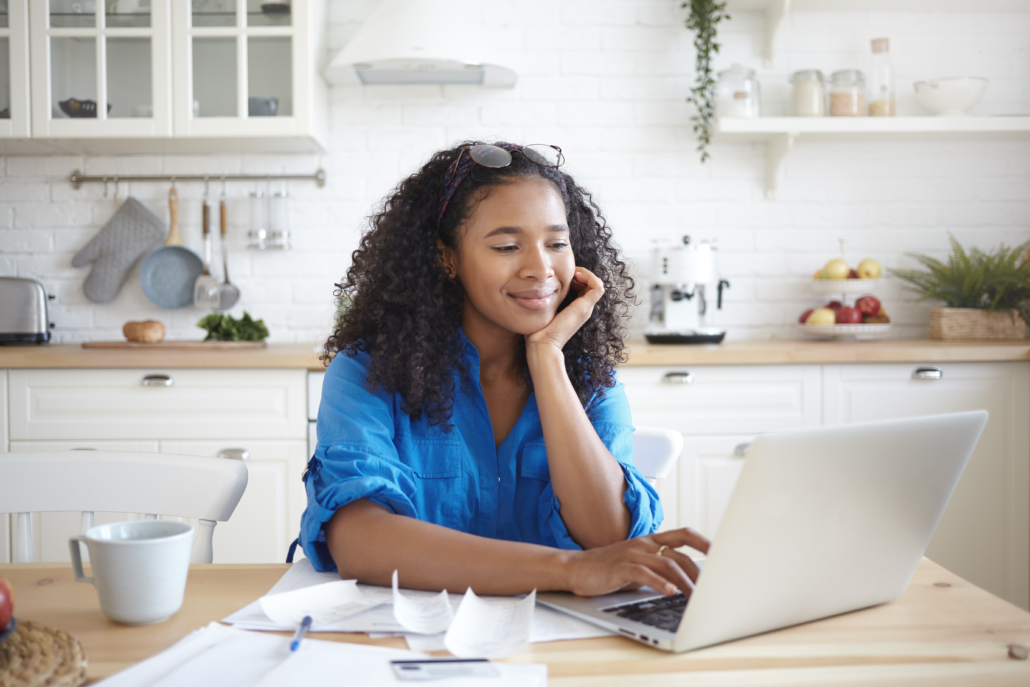 A happy young African American woman using a laptop computer and smiling, enjoying modern technology while paying bills for rent, gas and electricity online, sitting at kitchen table