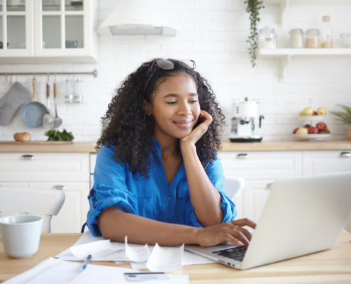 A happy young African American woman using a laptop computer and smiling on the road to financial wellness as a federal employee. Enjoying modern technology while paying bills for rent, gas and electricity online, sitting at kitchen table