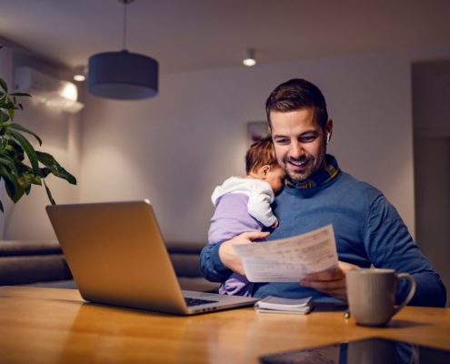 A man holds a sleeping baby while he reviews personal documents at home. Getting ahead on your finances for feds can set you and your family up for success