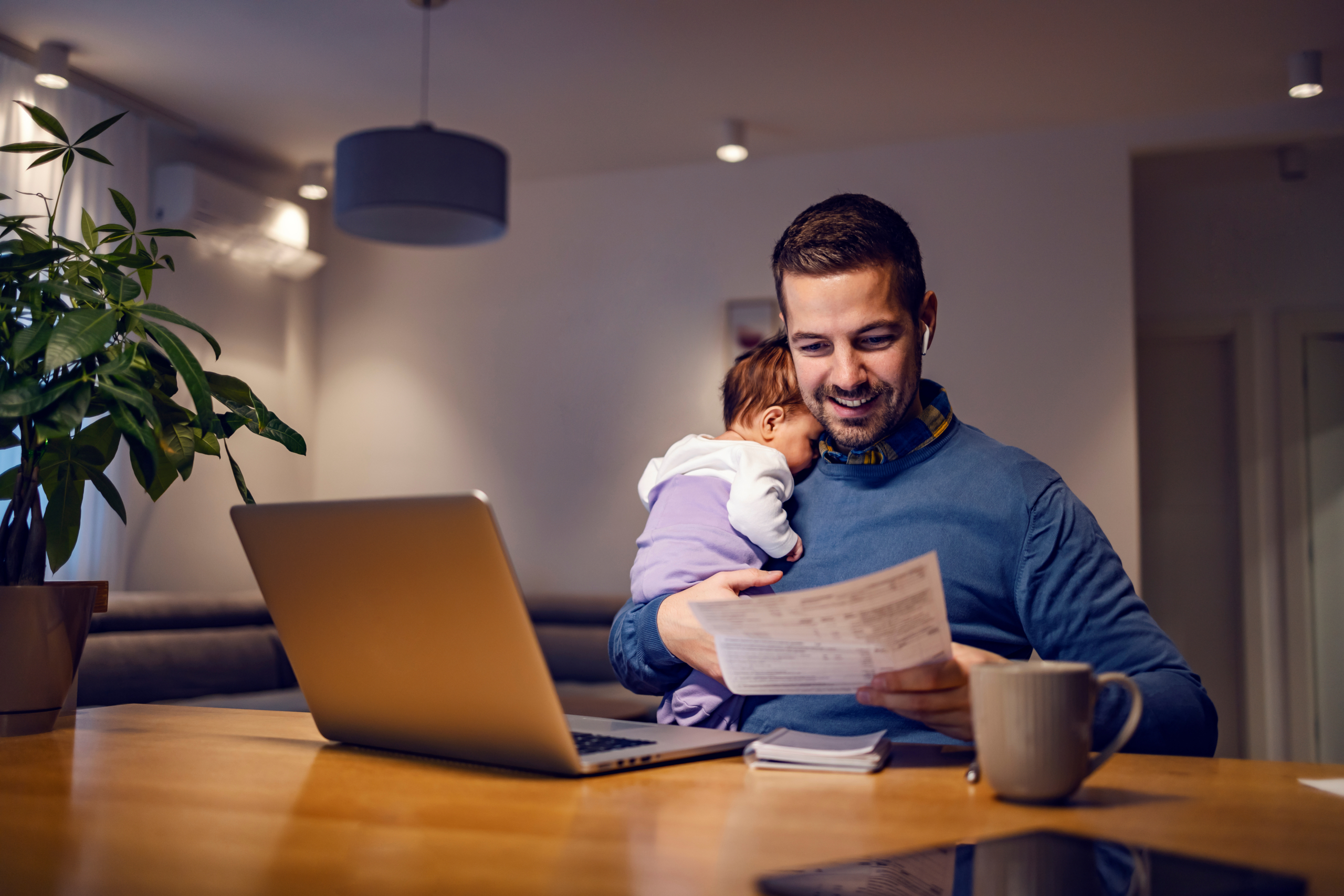 A man holds a sleeping baby while he reviews personal documents at home. Getting ahead on your finances for feds can set you and your family up for success